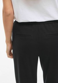 Juodos kelnės su lygiu paviršiumi, turinčios plokščią juosmens juostą, dvi galines kišenes ir tiesių kojų dizainą. Audinys atrodo minkštas ir lankstus.