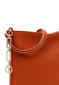 Orangefarbenes Leder-Handtäschchen mit glatter Textur, ausgestattet mit einem Schlaufenhenkel und goldfarbenen Beschlägen, darunter eine Kettenglieder und ein Akzentanhänger.