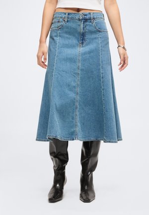 Femme portant une jupe en jean bleu mi-longue associée à des bottes en cuir noires montant jusqu'aux genoux, debout devant un fond uni.