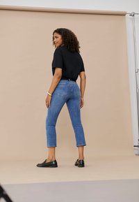 Femme aux cheveux bouclés portant un t-shirt noir, un jean bleu raccourci et des mocassins noirs, debout de profil sur un fond beige.