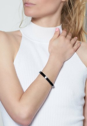 Schwarzes Metallarmband mit einem eleganten, flachen Design, das einen Kontrast zu dem weißen, gerippten ärmellosen Oberteil der Trägerin bildet. Glatte Oberfläche, minimaler Hardwareeinsatz.