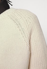 Stickad tröja i ljusbeige med texturerad väv, med en ribbad krage och accentstygn längs raglan ärmsömmarna.