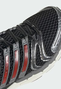 Chaussure de sport noire avec tige en mesh, lacets noirs, accents rouges sur les bandes latérales et semelle blanche texturée.