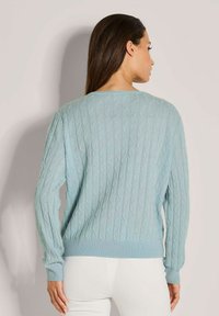 Eine Frau mit langen, dunklen Haaren trägt einen hellblauen, kabelgestrickten Pullover und weiße Hosen und steht vor einem hellen, einfarbigen Hintergrund.