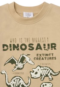 Beige T-Shirt mit schwarzen und grünen Dinosaurier-Skelettgrafiken und Text, Rundhalsausschnitt und kurzen Ärmeln.