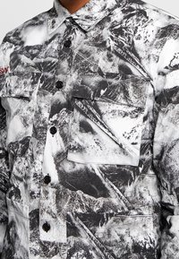 Giacca astratta in bianco e nero con motivo, con chiusura a bottoni e tasche a patta sul petto e in vita indossata da una persona.