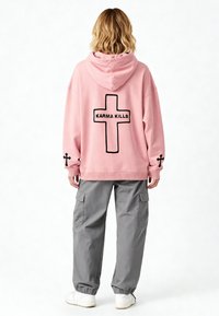 Felpa con cappuccio rosa con una croce nera e il testo "KARMA KILLS" sul retro; abbinata a pantaloni cargo grigi e sneakers bianche.