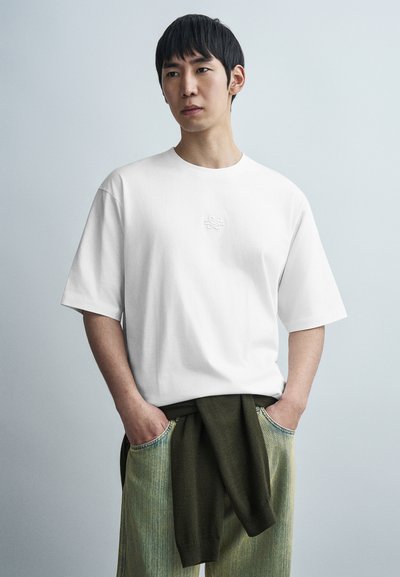 White cotton T-shirt with a round neck and short sleeves, featuring a subtle embossed logo on the chest. Paired with greenish denim pants.