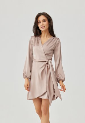 Kobieta o długich ciemnych włosach, ubrana w jedwabną beżową sukienkę kopertową z długimi bufkami, stojąca na tle gładkiego jasnoszarego tła.