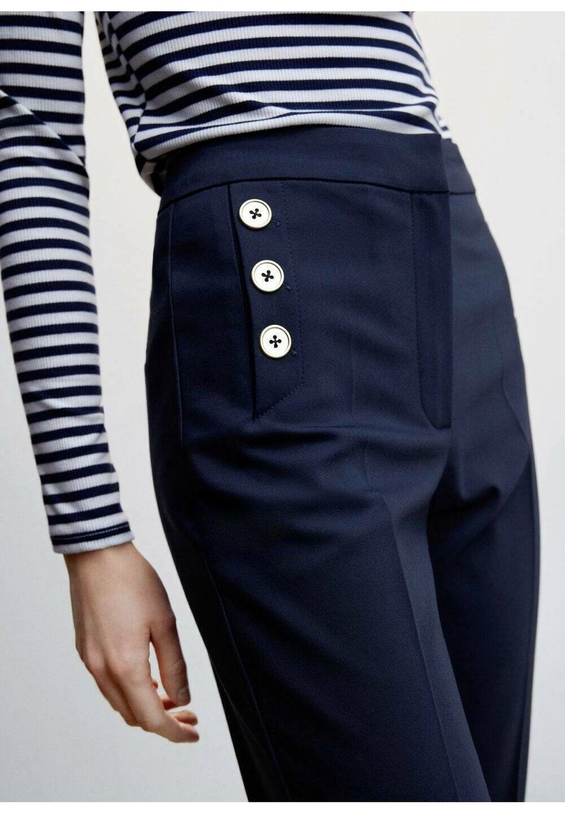 Pantalon bleu marine avec une texture lisse, doté de trois boutons blancs sur le côté et d'une coupe ajustée. Étendu sur un haut à rayures à manches longues.