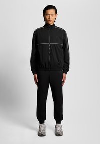 Άνδρας που κοιτάζει μπροστά φορώντας μαύρο αθλητικό μπουφάν με λευκές ρίγες, μαύρη φόρμα και γκρι αθλητικά παπούτσια, στέκεται μπροστά σε απλό γκρι φόντο.