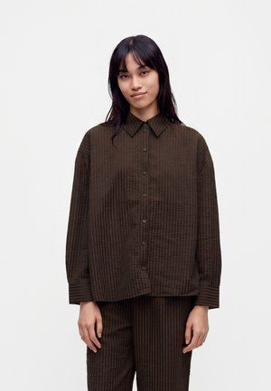 Jeune femme aux longs cheveux foncés portant une chemise ample marron foncé rayée à boutons et un pantalon assorti sur fond blanc uni.