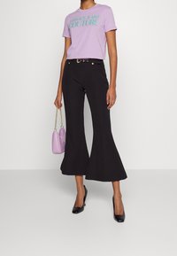 Kvinna i lavendelfärgad t-shirt från Versace Jeans Couture, svarta utsvängda byxor med bälte, svarta klackar, och hållande en lavendelfärgad handväska med guldkedja.