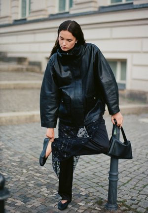 Une femme portant une veste en cuir noire pose un pied sur une borne tout en tenant une chaussure et un sac à main, dans une rue pavée.
