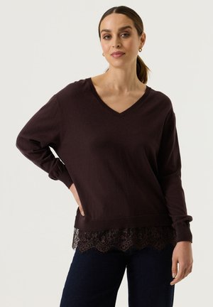 Vrouw met bruin haar samengebonden, die gouden oorringen draagt, een donkerbruine V-hals trui met kanten zoom en donkere broek, staand met hand op de heup.