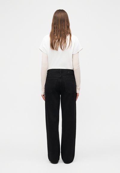 Γυναίκα με μακριά καστανά μαλλιά στέκεται με την πλάτη γυρισμένη, φοράει λευκό κοντομάνικο τοπ πάνω από μπεζ μακριά μανίκια και μαύρο παντελόνι με φαρδιά μπατζάκια.