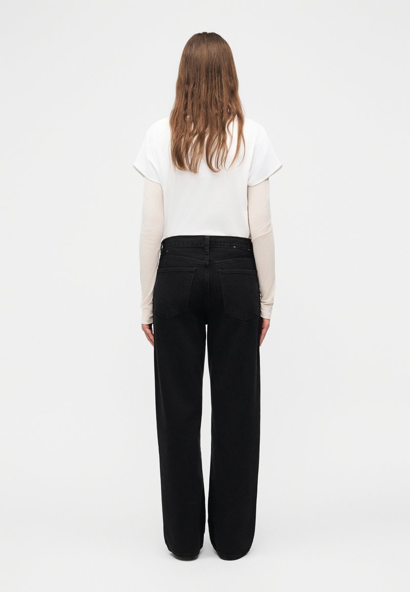 Femme aux longs cheveux bruns, vue de dos, portant un haut blanc à manches courtes sur un sous-pull beige à manches longues et un pantalon large noir.