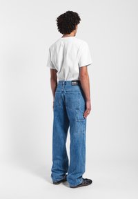 Pantalones cargo de mezclilla azul con un corte relajado, que cuentan con múltiples bolsillos y un parche de la marca en la parte trasera de la cintura. Combinado con una camiseta blanca.