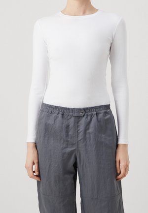 Osoba ubrana w dopasowany biały top z długim rękawem oraz luźne szare spodnie z elastycznym paskiem i zapięciem na guziki, stojąca na jednolitym tle.