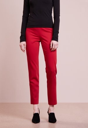 Kvinde iført røde slim-fit bukser, sort langærmet top og sorte hælede sko, stående mod en lys baggrund.