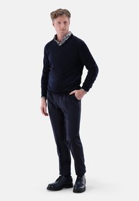 Marineblauer Pullover mit V-Ausschnitt, über einem gemusterten Hemd getragen; kombiniert mit dunklen Hosen und glänzenden schwarzen Schnürschuhen.