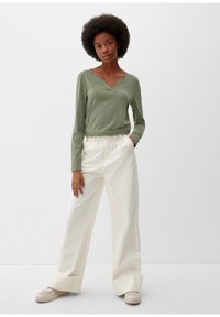 Olijfgroene top met lange mouwen en een V-hals ontwerp, gecombineerd met hoge witte wijde broek en beige sneakers, tegen een neutrale achtergrond.