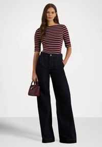 Žena v bordo-bielom pruhovanom topu a tmavých širokých džínsach s vysokým pásom drží malú bordovú kabelku, stojaca pred jednoduchým svetlým pozadím.