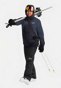 Skikleding bestaande uit een marineblauw jack, zwarte broek, handschoenen en witte skischoenen. De persoon draagt skistokken en een helm met een skibril.