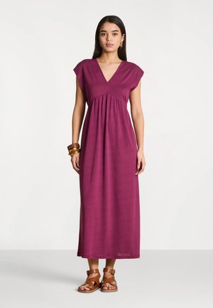 Femme debout portant une robe maxi violette sans manches, des sandales marron et des bracelets au poignet gauche sur un fond blanc uni.