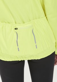 Neon gul lättviktsjacka med åtdragen midja, reflexdetaljer och en dragkedjeficka på baksidan. Slät textur, avslappnad design.
