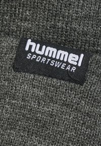 Tissu tricoté gris foncé avec une étiquette noire portant le texte blanc "hummel SPORTSWEAR". Surface texturée au toucher doux.
