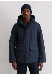 Marineblauwe waterdichte jas met capuchon, voorzien van twee zakken aan de voorkant en een ritssluiting. Gedragen over een gebreide trui, gecombineerd met een muts.