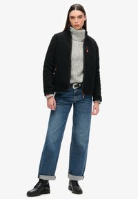 Zwarte fleece jas met een hoge kraag, grijze gebreide coltrui en blauwe vintage-stijl jeans, gecombineerd met zwarte veterschoenen.