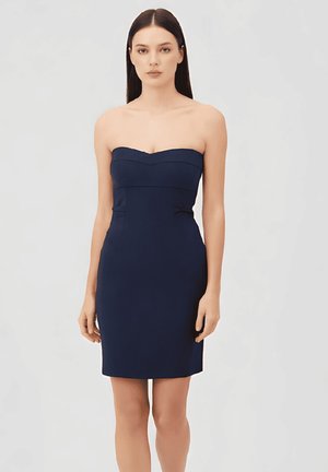 Mujer con cabello largo y oscuro, que lleva un vestido azul marino sin tirantes hasta la rodilla, de pie frente a un fondo claro y liso.