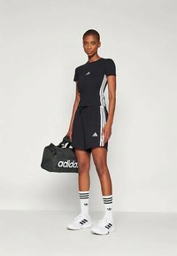 Schwarzes sportliches T-Shirt mit weißen Streifen, kombiniert mit einem passenden schwarzen Rock. Das Model hält eine schwarze Adidas-Duffle-Tasche und trägt weiße Sneaker.