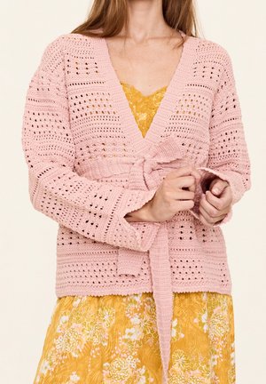 Femme portant un cardigan rose en crochet noué à la taille par-dessus une robe jaune à fleurs avec un détail en dentelle au niveau du décolleté.