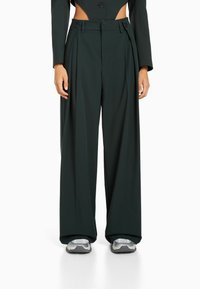 Mörkgröna, vida byxor med veck, två knappar vid midjan och en slät yta. Bärs med silvriga träningssneakers.