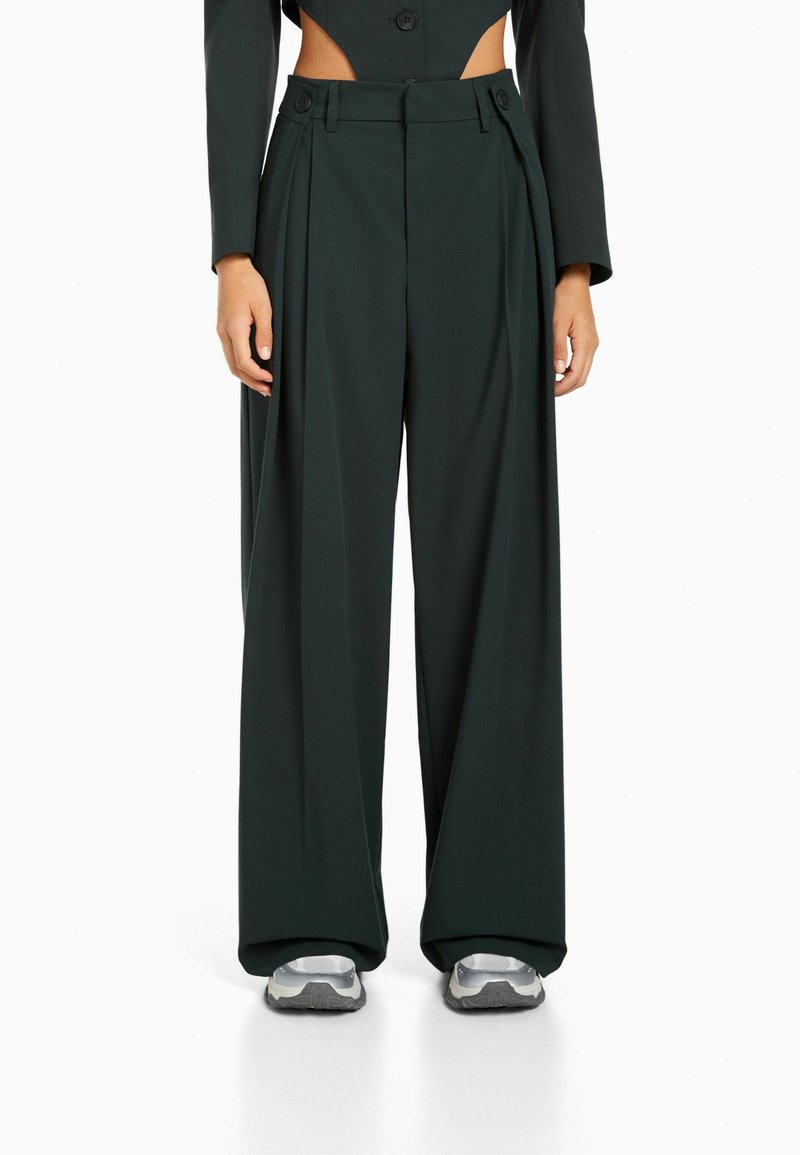 Mörkgröna, vida byxor med veck, två knappar vid midjan och en slät yta. Bärs med silvriga träningssneakers.