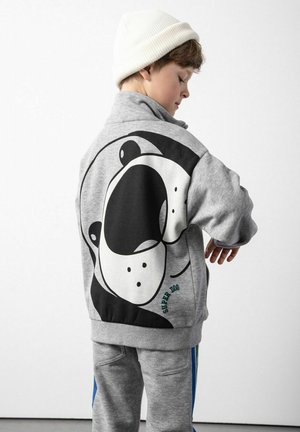 Szary sweatshirt z dużą czarno-białą grafiką psa na plecach, wykonany z materiału polarowego, z ściągaczami na rękawach. W zestawie ze szarymi spodniami.