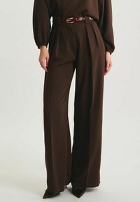 Pantalons larges marron avec des plis, fabriqués en tissu lisse. Assortis d'une ceinture slim bordeaux. Les pantalons ont une taille haute.