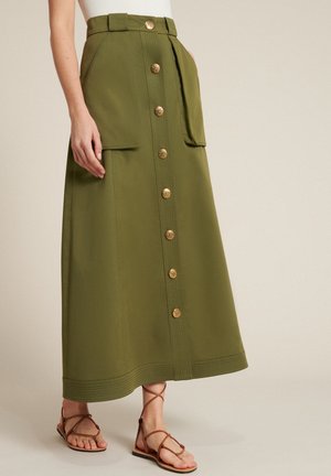 Femme portant une jupe longue vert olive avec des boutons dorés sur le devant, de grandes poches à l’avant et des sandales brunes à brides. Le haut du corps n’est pas visible.
