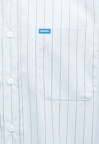 Λευκό πουκάμισο με λεπτές κάθετες μαύρες ρίγες, τσέπη μπροστά δεξιά, μπλε ετικέτα με την ένδειξη "HUGO" και λευκά κουμπιά στην αριστερή πλευρά.