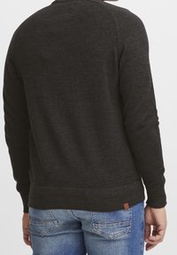 Pull gris foncé en tissu tricoté doux, avec un col rond, un ourlet côtelé et une petite étiquette logo marron à l'arrière près de l'ourlet.