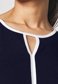 Tmavě modrý top s bílým výstřihem a klíčovým otvorem na hrudi. Vyrobený z elastické látky s hladkou texturou.