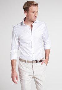 Camicia bianca a maniche lunghe con colletto a punta e chiusura con bottoni, abbinata a pantaloni in lino di colore chiaro e una cintura intrecciata.