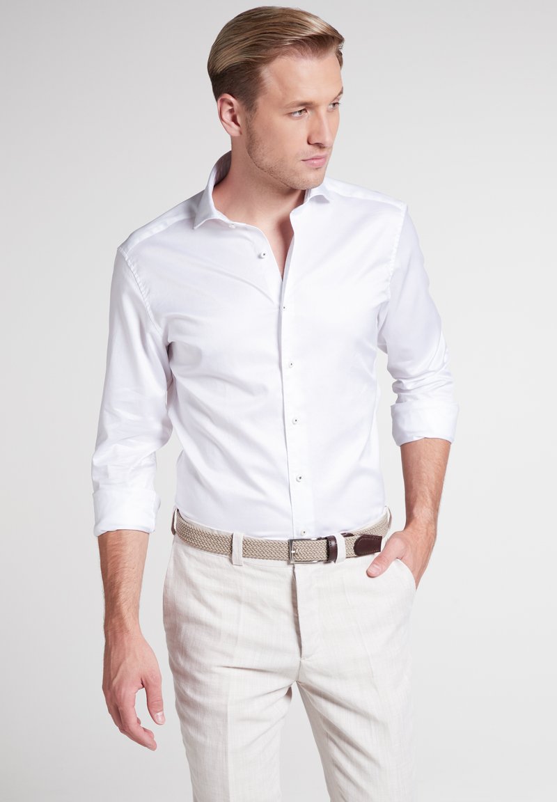 Camicia bianca a maniche lunghe con colletto a punta e chiusura con bottoni, abbinata a pantaloni in lino di colore chiaro e una cintura intrecciata.