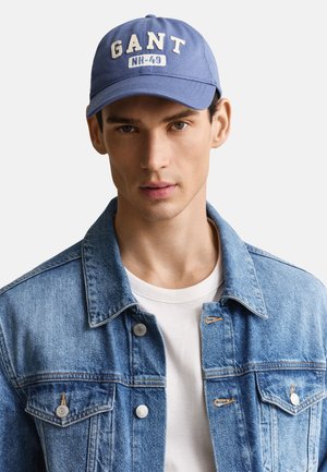 Jonge man die een blauwe baseballpet draagt met de tekst "GANT NH-49", een spijkerjack over een wit t-shirt, die recht vooruit kijkt tegen een eenvoudige achtergrond.