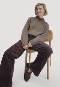Bruine gebreide trui met een hoge hals, gecombineerd met donkerpaarse corduroy wijde broek. Model zit op een licht houten stoel en draagt bruine schoenen.