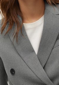 Femme portant un blazer à double boutonnage à motif pied-de-poule gris et noir sur un t-shirt blanc à col rond, avec des cheveux bruns visibles sur un côté.