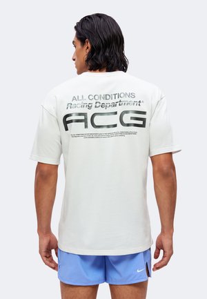 Uomo con capelli scuri che indossa una maglietta bianca a maniche corte con la scritta "ALL CONDITIONS Racing Department ACG" e pantaloncini Nike azzurri chiaro.